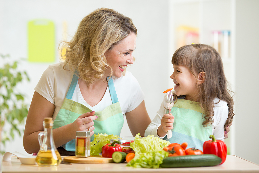 Mujer blanca de cabello claro usando delantal menta y camiseta blanca mira a su hija de camibuso blanco y el mismo delantal, de cabello largo color café mientras sostiene un tenedor en la mano derecha. Frente a ellas una mesa con alimentos saludables.