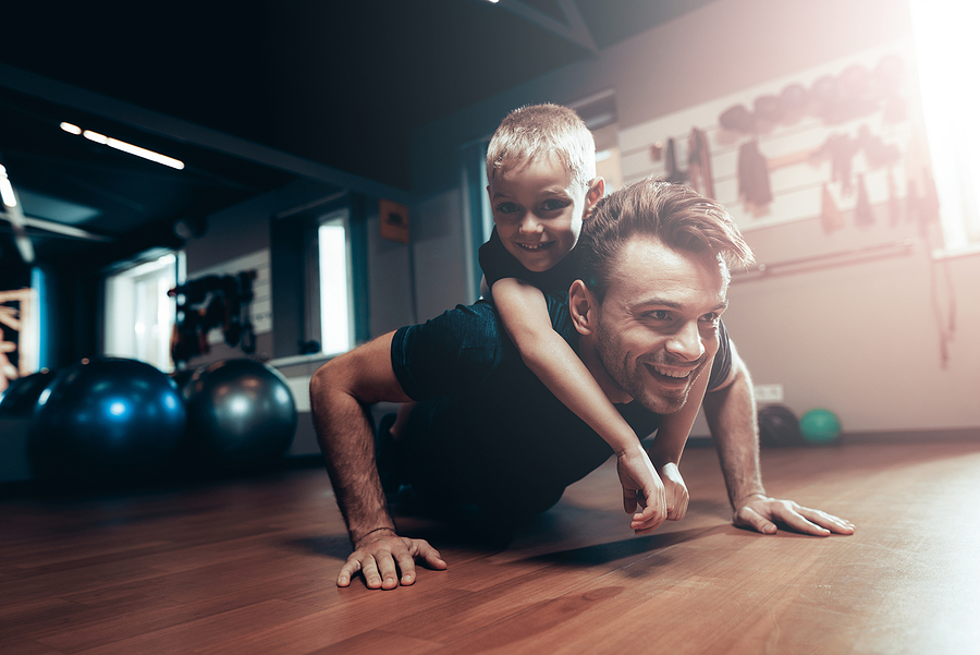 En un gimnasio papá e hijo hacen ejercicio. Hombre blanco de cabello castaño, adulto con camiseta negra y sobre él, abrazado un niño de cabello con camiseta negra